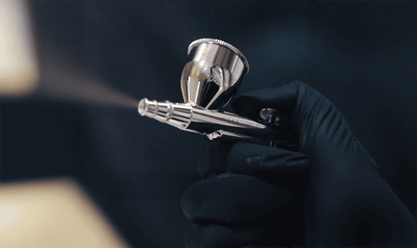 A hand wearing a black glove holds an airbrush, spraying a fine mist against a dark background—an image reminiscent of precision treatments at a klinika medycyny estetycznej.