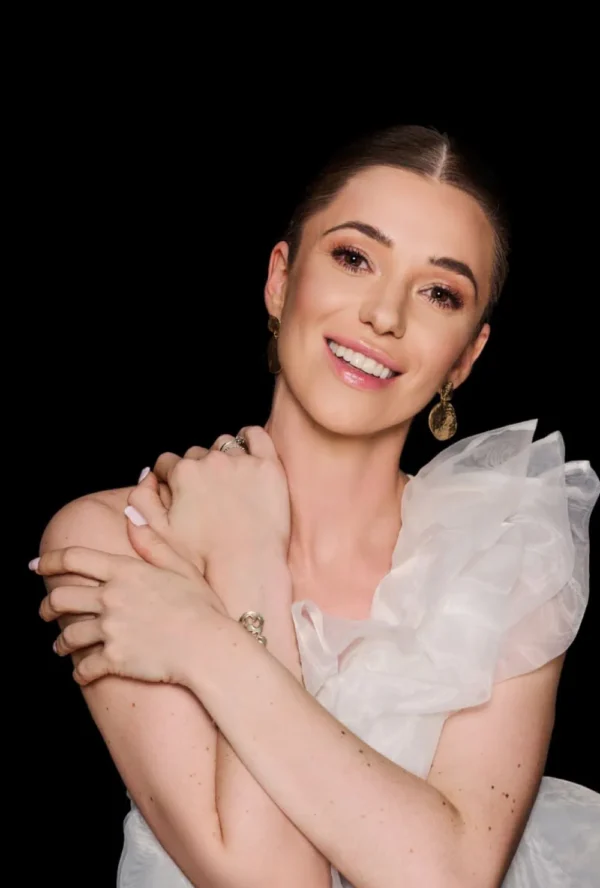 Uśmiechnięta kobieta z lekkim makijażem i ułożonymi brązowymi włosami pozuje na czarnym tle. Ukazując naturalne piękno, podkreśla pewność siebie, jaką może wzbudzić medycyna estetyczna Wrocław, ubrana w białą sukienkę z odkrytymi ramionami i złote kolczyki.