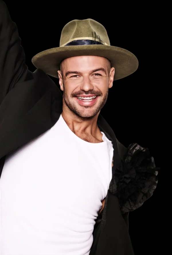 Uśmiechnięta osoba w kapeluszu z szerokim rondem, białej koszuli i czarnej kurtce pozuje na jednolitym czarnym tle, uosabiając pewność siebie i odświeżony wygląd często poszukiwany przez medycynę estetyczną Wrocław.