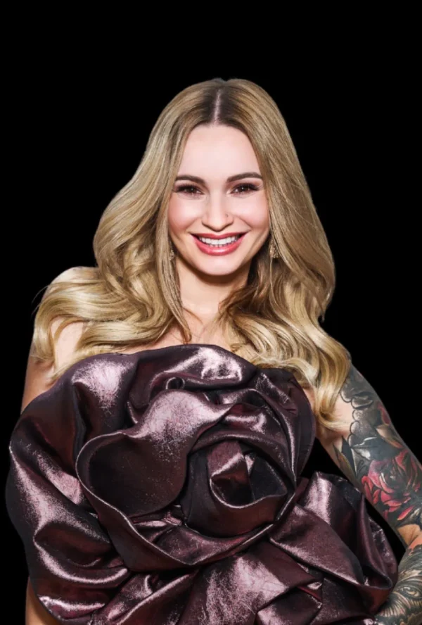 Uśmiechnięta kobieta z długimi blond włosami ma na sobie dużą, metaliczną sukienkę w kształcie róży i pokazuje swój kolorowy rękaw tatuażu - jej wygląd jest tak odważny, jak najnowsze trendy w medycynie estetycznej Wrocław - pozując na czarnym tle.