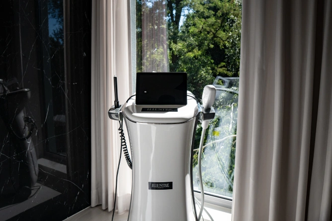 Eleganckie białe urządzenie medyczne lub kosmetyczne używane w medycynie estetycznej we Wrocławiu stoi przy oknie z białymi zasłonami, odbijając drzewa i światło słoneczne z zewnątrz.