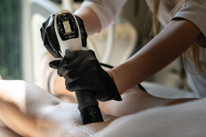 Osoba w czarnych rękawiczkach używa ręcznego urządzenia medycznego lub kosmetycznego na udzie klienta, prawdopodobnie wykonując nieinwazyjny zabieg modelowania sylwetki w klinice medycyny estetycznej we Wrocławiu.