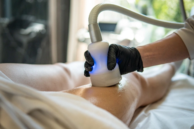 Osoba poddaje się zabiegowi kosmetycznemu na nodze za pomocą ręcznego urządzenia emitującego niebieskie światło, obsługiwanego przez osobę w czarnych rękawiczkach w otoczeniu przypominającym spa, prezentując zaawansowaną medycynę estetyczną Wrocław.