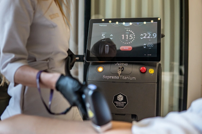 Epilacja laserowa twarzy Wrocław – skuteczne usuwanie owłosienia w delikatnych partiach Osoba w białym fartuchu korzysta z urządzenia do depilacji laserowej Soprano Titanium, często spotykanego w klinikach medycyny estetycznej we Wrocławiu. Operator trzyma rękojeść urządzenia podczas leczenia pacjenta, a ekran dotykowy wyświetla ustawienia i dane.Epilacja laserowa twarzy Wrocław