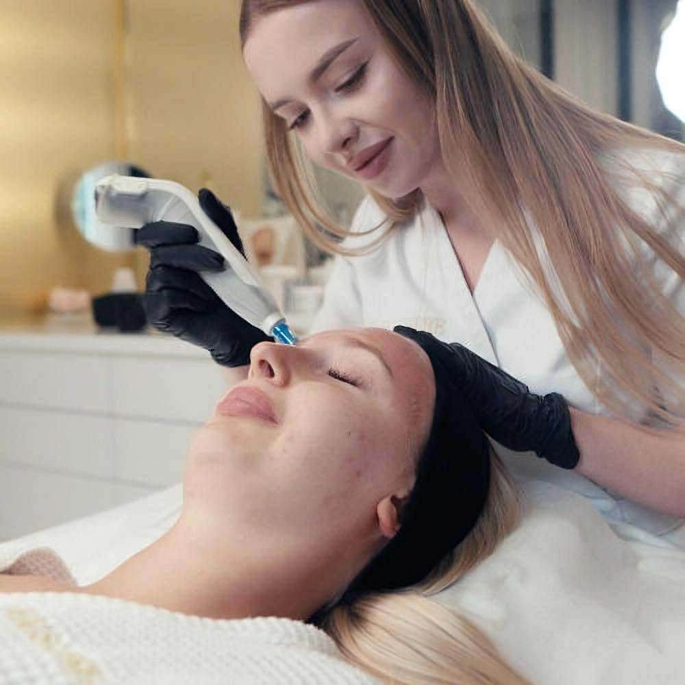 A skincare professional wearing black gloves performs a facial treatment with a handheld device on a client’s face in a modern, well-lit klinika medycyny estetycznej. The client lies relaxed with eyes closed.