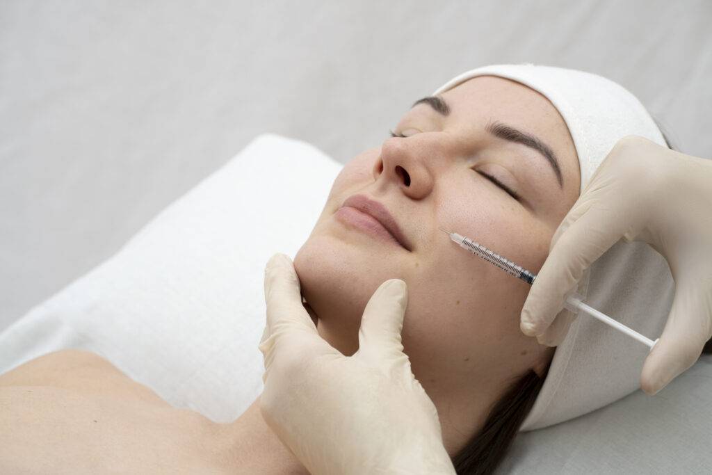 A woman with closed eyes lies on a treatment bed while a gloved medical professional injects her cheek with a syringe at a klinika medycyny estetycznej, specializing in medycyna estetyczna Wrocław.