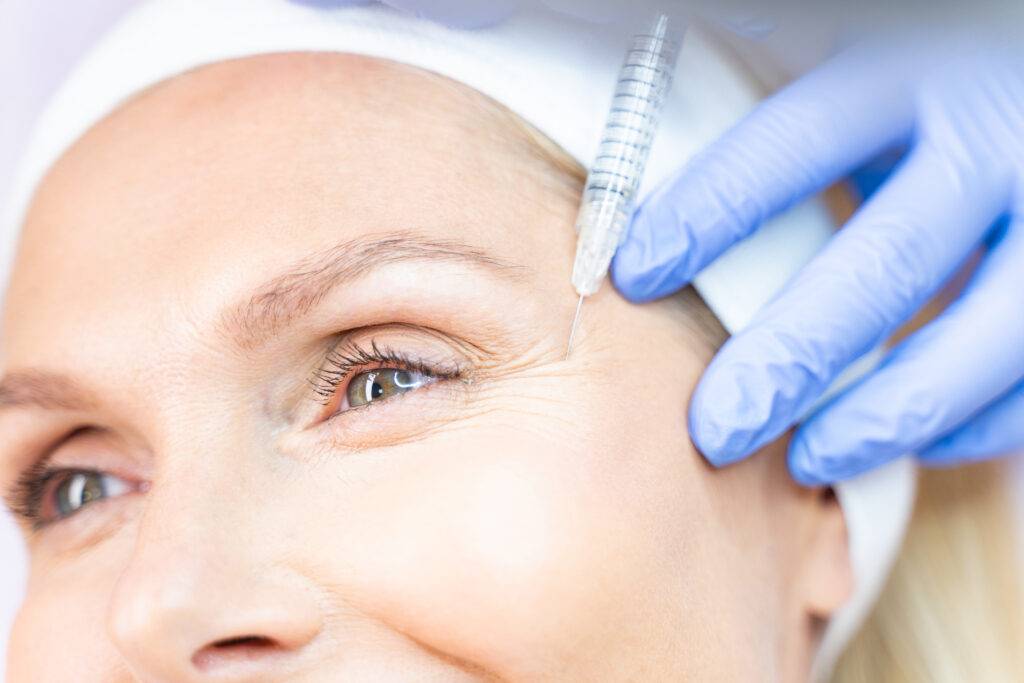 A close-up of a woman's face as a medical professional from a klinika medycyny estetycznej, wearing blue gloves, injects a syringe near her eye—possibly for a cosmetic procedure like Botox.