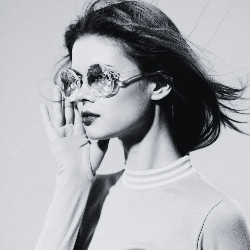A woman with shoulder-length hair wears large, crystal-like sunglasses and a high-neck top. Posing with one hand near her face, she looks to the side—her style reminiscent of a klinika medycyny estetycznej photo shoot. The image is in black and white.