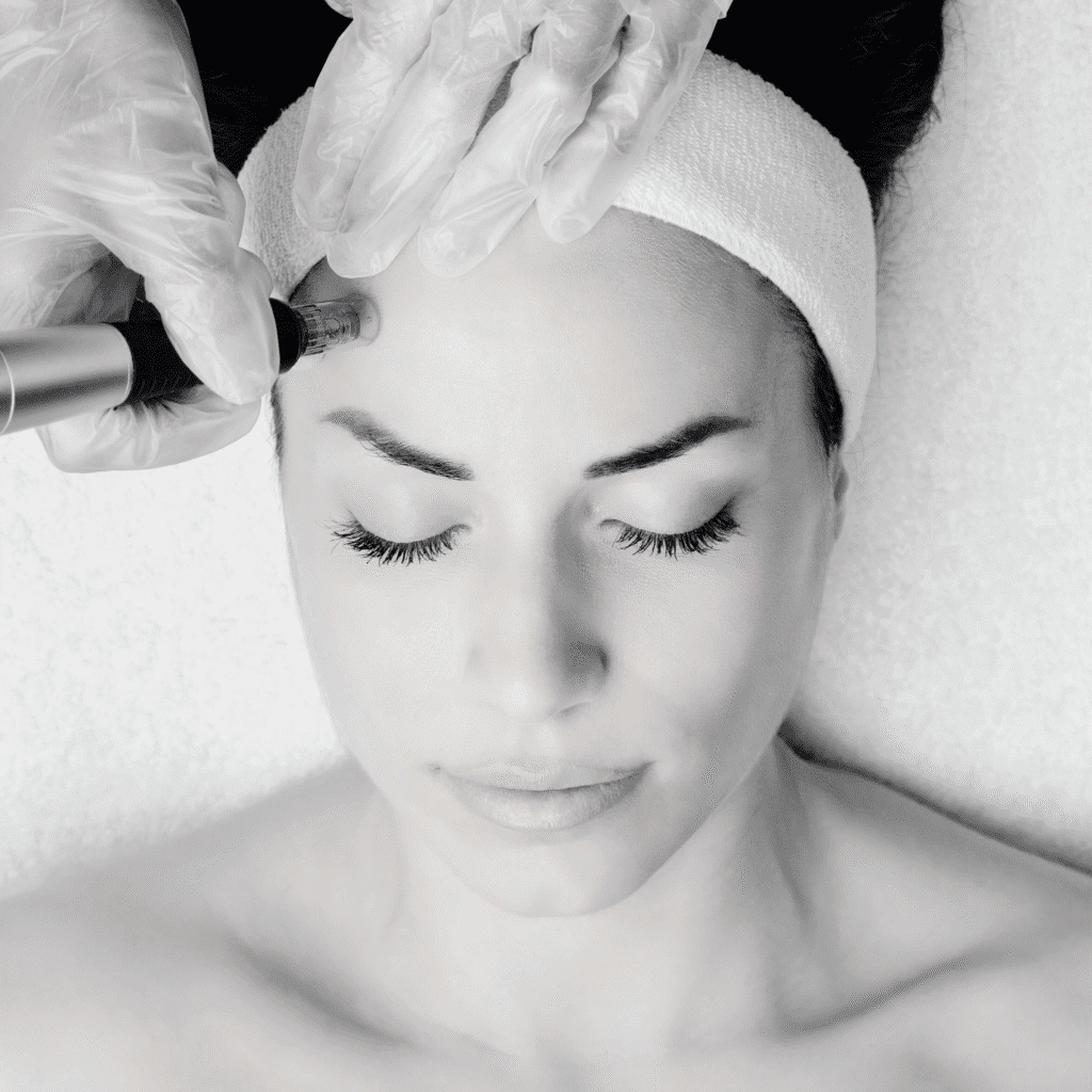 A woman with closed eyes lies on a treatment bed, wearing a white headband, while a gloved professional at a klinika medycyny estetycznej performs a skincare procedure on her forehead using a handheld device.