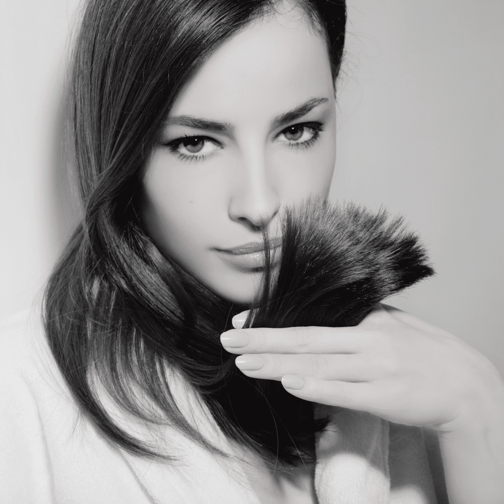 A woman with straight dark hair and intense eyes holds a section of her hair in front of her face, inspecting the ends. The black-and-white image highlights her smooth hair and contemplative look, echoing the precision of medycyna estetyczna Wrocław.