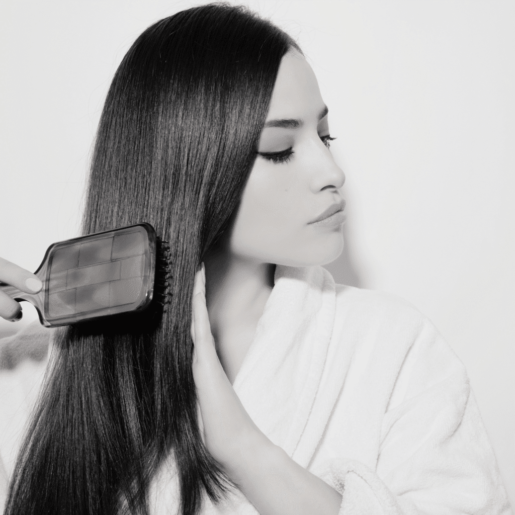 A woman in a white robe brushes her long, straight, dark hair while looking to the side against a plain background, evoking the serene atmosphere of a klinika medycyny estetycznej.