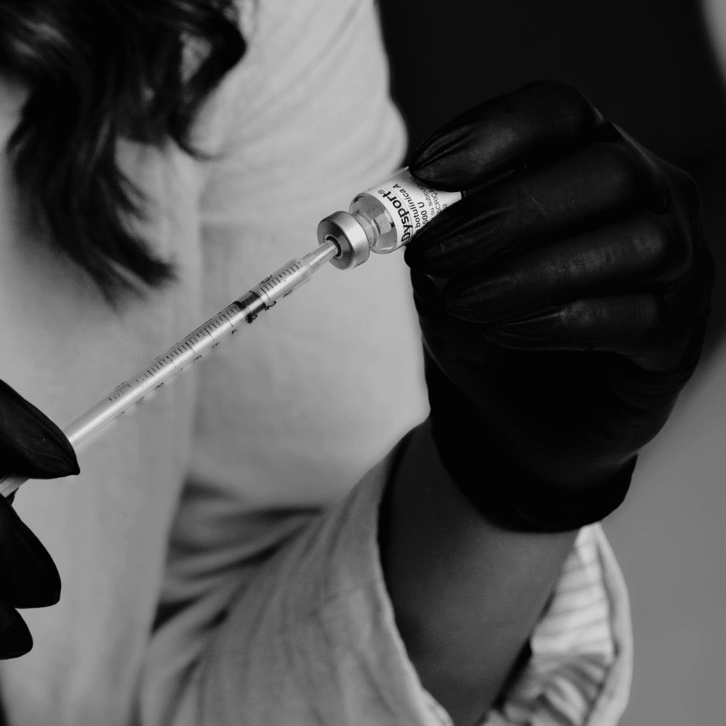 Wolumetria twarzy A person wearing black gloves fills a syringe with liquid from a small vial, preparing for an injection—a scene often found in klinika medycyny estetycznej or during medycyna estetyczna Wrocław procedures. The black and white image shows only the hands and part of a lab coat.