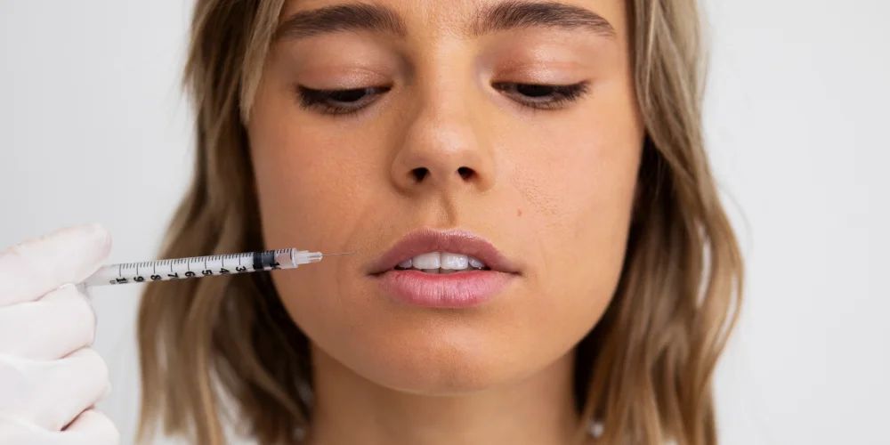 A person with light brown hair looks down as a gloved hand holds a syringe near their lips, preparing for an injection—powiększanie ust Wrocław offered at a klinika medycyny estetycznej. The background is plain and white.