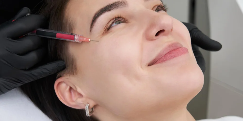 A woman lies on her back, smiling, as a gloved hand injects a syringe with red liquid near her eye during a medycyna estetyczna Wrocław procedure at a klinika medycyny estetycznej.