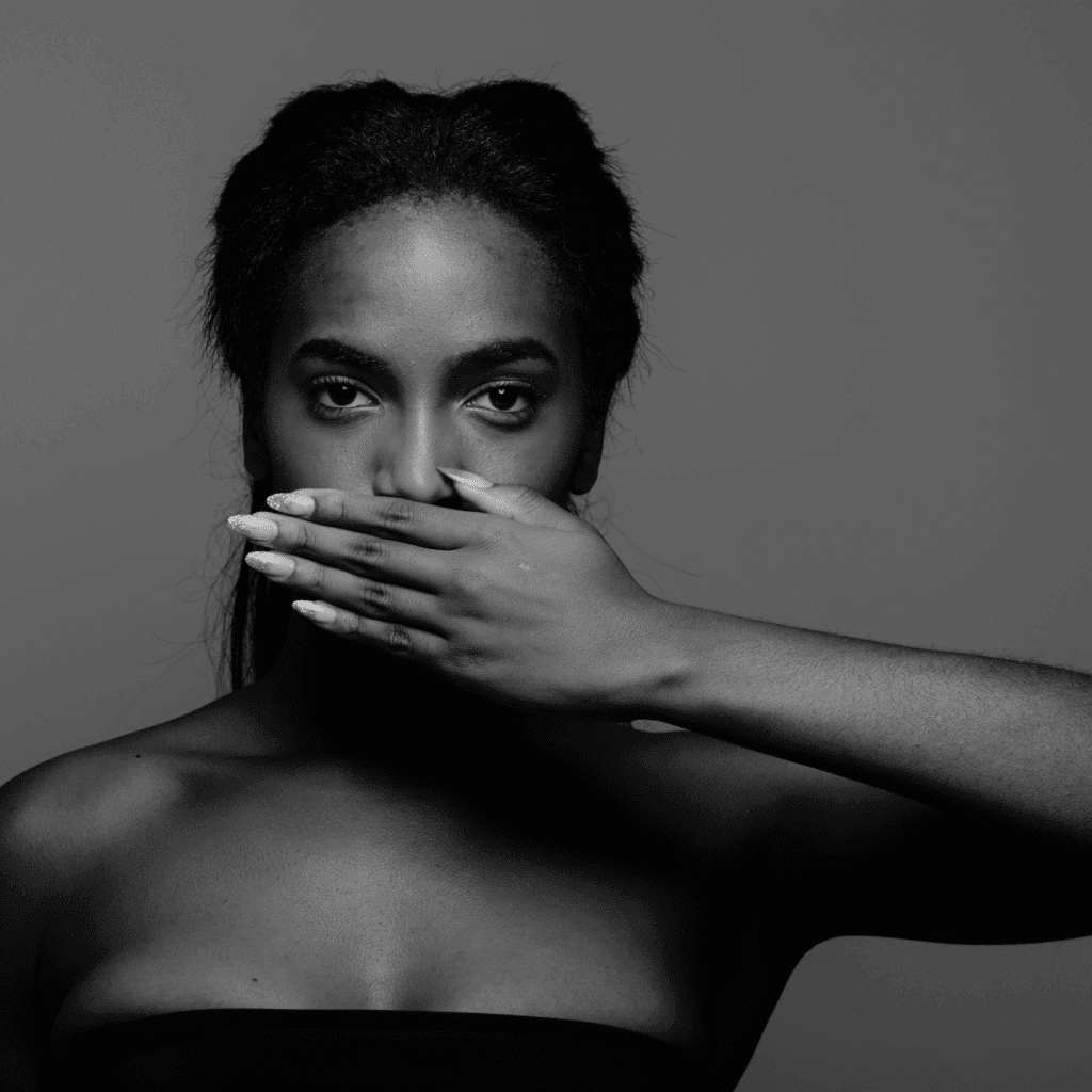 A woman in a strapless top holds her hand over her mouth, looking directly at the camera against a plain gray background. The black and white image is perfect for illustrating powiększanie ust Wrocław at a klinika medycyny estetycznej.