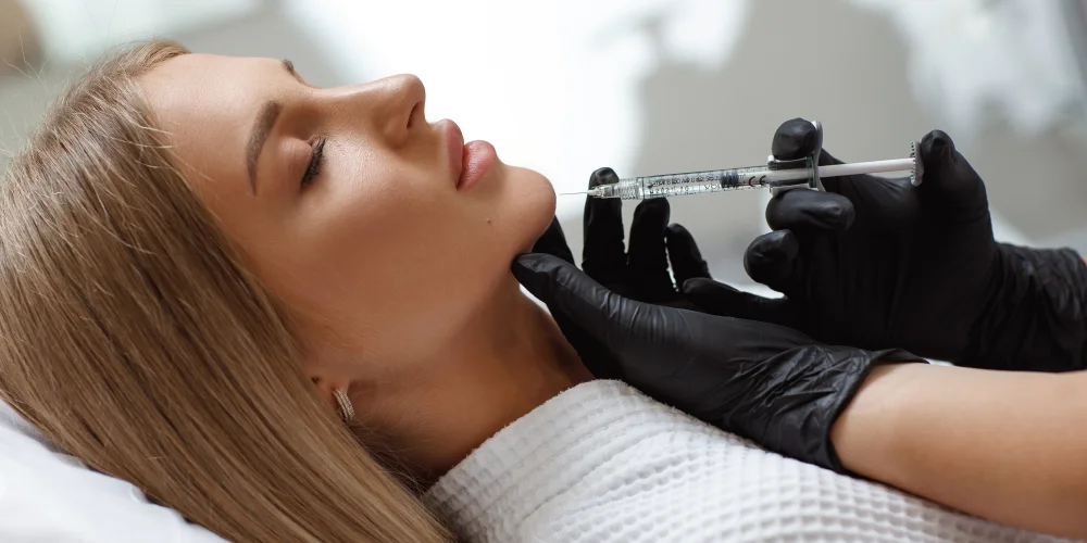 A woman with long blonde hair lies calmly in a white robe as a professional at a klinika medycyny estetycznej injects her chin, likely during a medycyna estetyczna Wrocław cosmetic procedure.