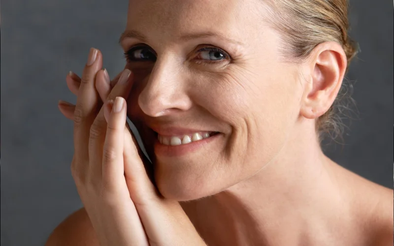 A smiling woman with light hair pulled back holds her hands and a phone to her face, looking directly at the camera against a neutral background—perfect for a klinika medycyny estetycznej or medycyna estetyczna Wrocław promotion.