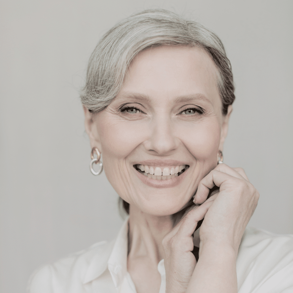 A smiling older woman with gray hair styled back, wearing hoop earrings and a white blouse, poses with one hand near her chin against a light background—radiating confidence and beauty after visiting klinika medycyny estetycznej.