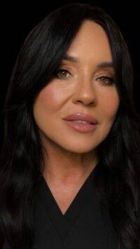 A woman with long, dark wavy hair and light makeup looks at the camera with a neutral expression. She is wearing a dark top; the photo has a black background, evoking the elegance often seen in medycyna estetyczna Wrocław clinics.